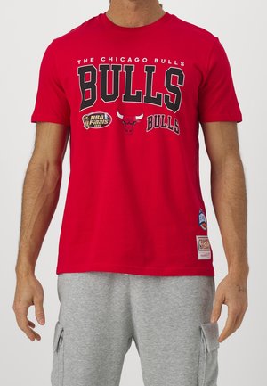 Mann trägt rotes Chicago Bulls T-Shirt mit NBA Finals 1996 Aufnäher und graue Cargoshorts, steht vor weißem Hintergrund.
