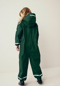 Grön vattentät regndräkt med huva, elastiska ärmslut och öppningar vid anklarna. Har vitt accentmaterial och en dragsko i midjan för en figursydd look.