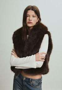 Hnědá vesta z umělé kožešiny se širokým límcem, oblečená přes bílý top s dlouhým rukávem. Modelka má rovné tmavé vlasy a zlatý prsten na ruce.