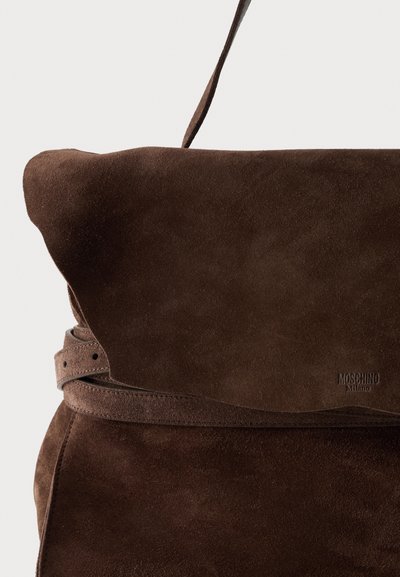 Καφέ σουέντ τσάντα με επάνω πτερύγιο και λεπτό ρυθμιζόμενο λουράκι, με ανάγλυφο λογότυπο "Moschino Milano" στην κάτω δεξιά γωνία.