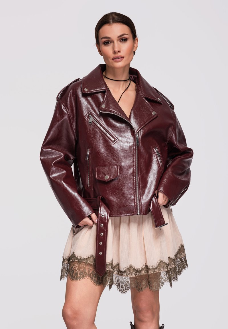 Femme portant une veste en cuir bordeaux brillante sur une jupe beige bordée de dentelle, debout les mains dans les poches de la veste, sur un fond uni.