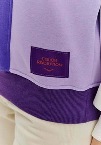 Fioletowa bluza z przeskokiem, z ribbowanym wykończeniem, w połączeniu jasnych i ciemnych odcieni fioletu oraz naszywką z napisem "COLOR REVOLUTION" w kolorze pomarańczowym.