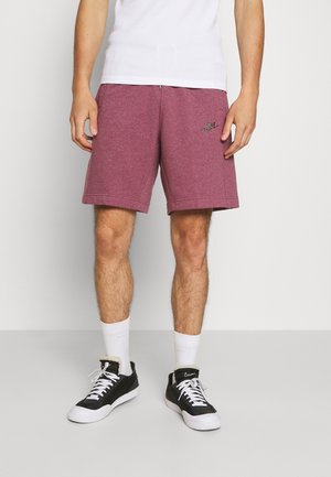 Man in een paarse Nike-shorts, witte sokken, zwart-witte Nike-sneakers en een wit T-shirt, staande tegen een effen lichte achtergrond.