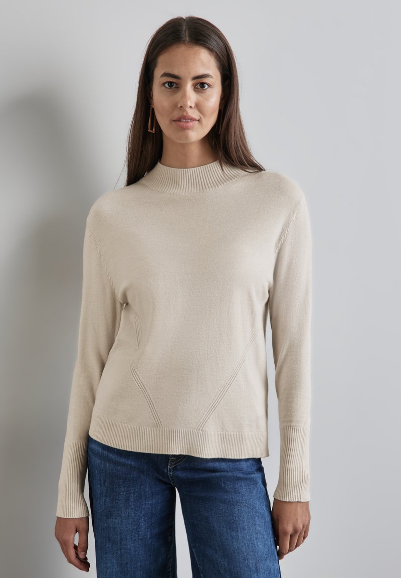 Beige, strukturierter Pullover mit hohem Rippkragen, seitlichen Nähten und langen Ärmeln, kombiniert mit blauen Jeans.