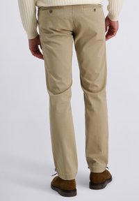 Mann in beigen Chinos, braunen Wildlederschuhen und einem cremefarbenen Pullover, der vor einem schlichten, hellen Hintergrund steht.