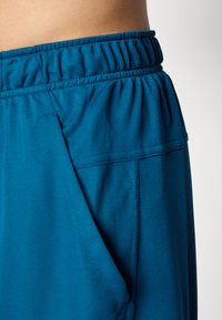 Close-up of blue athletic shorts with elastic waistband and side pocket on a person's waist against a plain background.