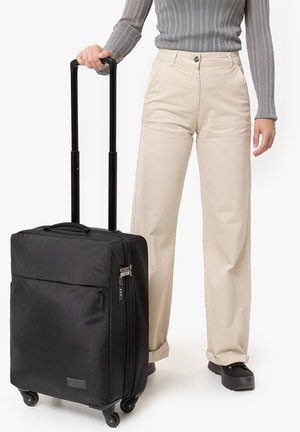 Black wheeled suitcase with a rectangular shape, fabric material, top handle, zippered pocket, and four wheels, positioned beside light-colored pants.