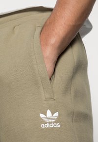 Ljus olivgröna joggers gjorda av mjukt material, med sidofickor och en vit broderad Adidas-logotyp framtill.