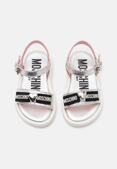 Sandales métalliques argentées avec des sangles élastiques noires arborant la marque "Moschino" et un détail en forme de cœur. Semelles blanches et doublure intérieure rose.