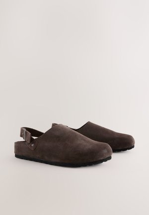 Zoccoli in camoscio marrone con punta arrotondata, cinturino posteriore e suola testurizzata. Presentano una finitura liscia e un design minimalista.