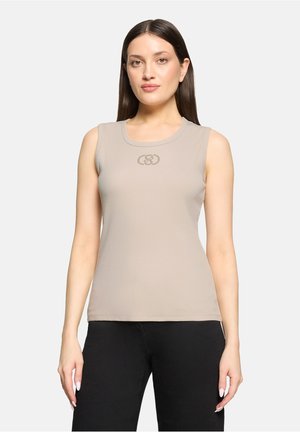 Femme aux cheveux raides et foncés portant un haut beige sans manches avec un petit motif circulaire sur la poitrine et un pantalon noir, debout devant un fond blanc.