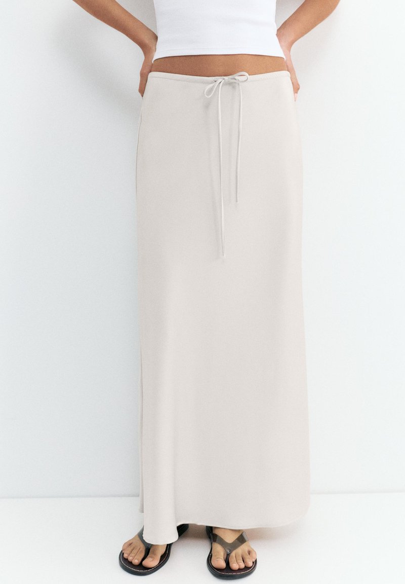 Femme portant une jupe longue beige à cordon, un débardeur court blanc et des tongs noires, debout devant un fond blanc.