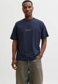 Tume sinine puuvillane T-särk lühikeste varrukatega, millel on kuldne käekirjaga tekst "Original Studio" ees, koos heledate teksadega.