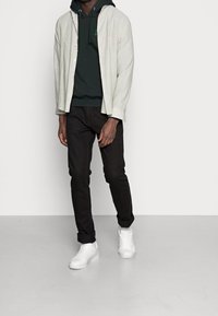 Camicia button-up grigia chiara sopra un felpa con cappuccio verde scuro, abbinata a jeans neri e sneakers bianche, che mostrano un look casual a strati.