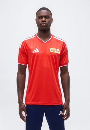 Mand stående fremadrettet iført rød 1. FC Union Berlin fodboldtrøje og mørkeblå Adidas bukser mod ensfarvet baggrund.