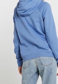 Ljusblå hoodie i bomullsblandning med huva, ribbade ärmmuddar och nederkant, i en avslappnad passform som framhävs av den slitna denimbakgrunden.