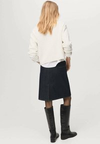 Maglione cropped a trecce bianche sopra una gonna A-line in denim scuro, abbinato a stivali neri fino al ginocchio. Texture liscia e linee pulite visibili.