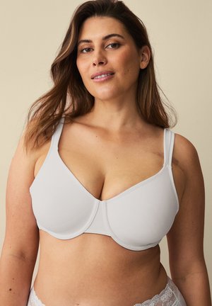 Frau mit mittellangem braunem Haar, die einen hellgrauen stützenden BH und-Unterwäsche mit Spitzenbesatz trägt und vor einem beigen Hintergrund steht.