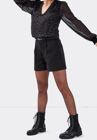 Blouse noire transparente à manches longues avec un col en V et des points scintillants, assortie à un short noir taille haute et des bottines à lacets noires.