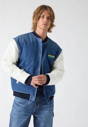 Young man with shoulder-length hair wears a blue and white varsity jacket and blue jeans, standing against a plain light background.