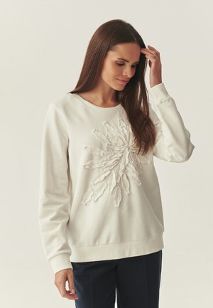 Vrouw met lang bruin haar draagt een witte trui met reliëfbloemenpatroon, raakt haar haar aan en staat tegen een effen lichte achtergrond.