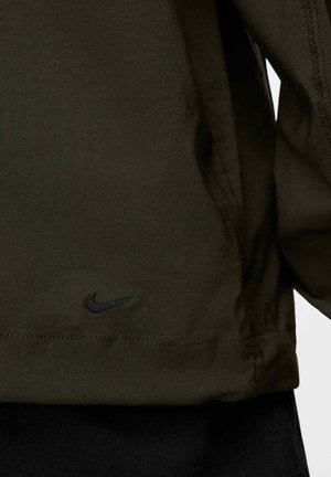 Chaqueta Nike de tela verde oscuro con una textura suave; presenta un logo negro bordado en el dobladillo y detalles de costura en contraste.