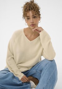 Pull en tricot beige avec un col en V et une texture côtelée. Porté avec un jean large bleu clair et des chaussures marron, pose assise.