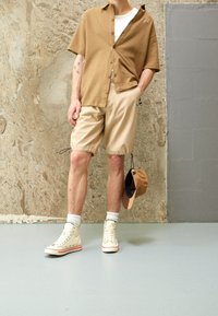 Chemise courte à manches courtes de couleur brun clair, shorts beiges, baskets montantes crème avec des accents rouges, tenant une casquette marron ; fond de mur lisse.