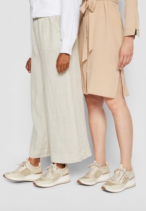 To kvinder iført beige sneakers; den ene med brede lyse beige bukser og hvid skjorte, den anden med en beige kjole med sideslids.