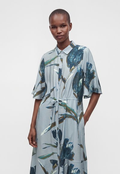 Femme aux cheveux courts porte une robe bleu clair avec de grands imprimés floraux bleu foncé et verts, nouée à l'avant, manches courtes et un col.