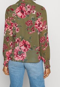 Blusa floreale verde oliva con fiori rosa e rossi. Maniche lunghe con polsini elastici. Tessuto leggero, leggermente trasparente, vestibilità comoda.