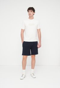 Άσπρο βαμβακερό t-shirt με διακριτικό γραφικό σχέδιο, συνδυασμένο με σκούρα μπλε σορτς. Το μοντέλο φοράει λευκά αθλητικά παπούτσια και κάλτσες μήκους μέχρι τον αστράγαλο.