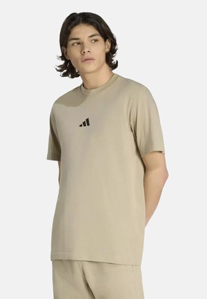 Giovane uomo con capelli di media lunghezza che indossa una t-shirt beige Adidas e pantaloncini abbinati, che guarda di lato su uno sfondo uniforme.