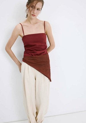 Woman with light skin and brown hair wears a sleeveless burgundy top and loose cream trousers, standing against a plain white wall.