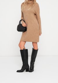 Bottes noires montantes jusqu'au genou avec un bout carré, un talon bloc, des fermetures éclair latérales et des languettes de tirage. Portées avec une robe beige en maille et un sac à main noir.