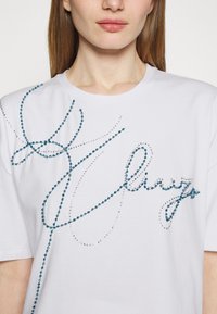 Camiseta de algodón blanca con rhinestones azules y transparentes dispuestos en un diseño en cursiva en la parte delantera. Mangas cortas y escote redondo.