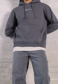 Felpa con cappuccio grigia in pile con tasca frontale e logo bianco, indossata sopra una camicia color crema, abbinata a pantaloni in denim grigi. Tessuto strutturato, vestibilità comoda.