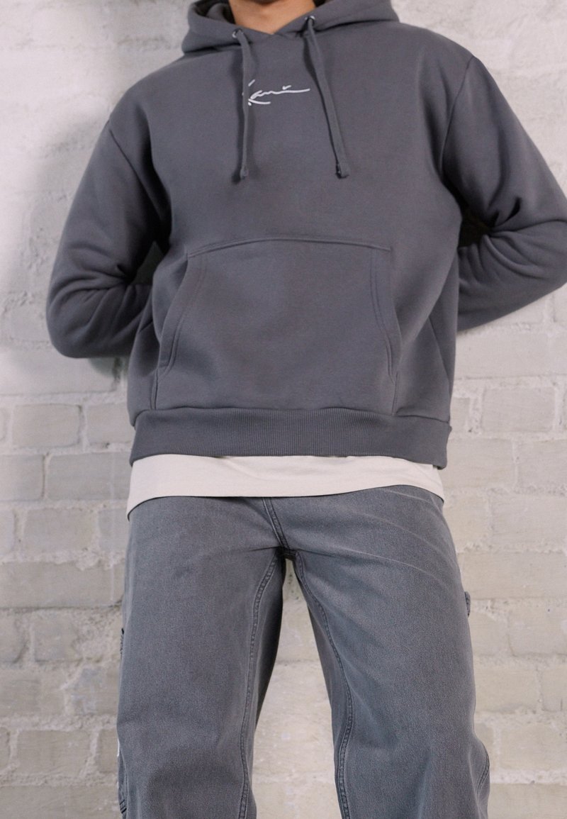 Felpa con cappuccio grigia in pile con tasca frontale e logo bianco, indossata sopra una camicia color crema, abbinata a pantaloni in denim grigi. Tessuto strutturato, vestibilità comoda.