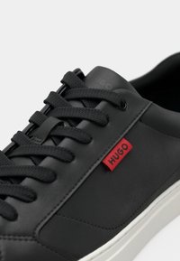 Zwarte leren sneaker met een gestructureerd oppervlak, ronde neus en witte rubberen zool. Voorzien van zwarte veters en een rode "HUGO" logo-label aan de zijkant.