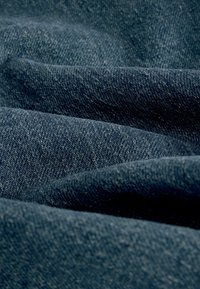 Denim-kangas tummansinisessä värissä, jossa on teksturoitu pinta, jossa on hienovaraisia sävyerotuksia ja tiheä kudontamalli, näyttäen taitteita ja ryppyjä.