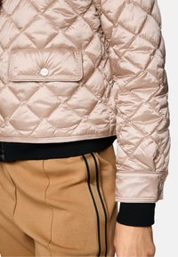 Gepolstete pinke Pufferjacke mit einer Druckknopfklappe und schwarzen Rippstrickbündchen, kombiniert mit hellbraunen Hosen mit schwarzen Seitenstreifen.