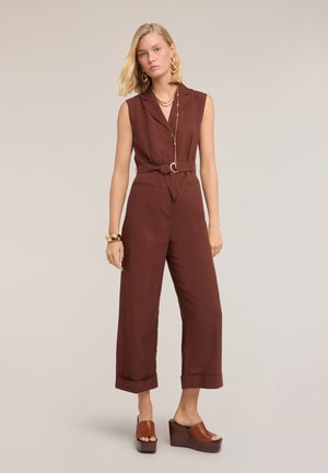 Kobieta w brązowym kombinezonie bez rękawów z paskiem, szerokimi nogawkami oraz brązowych sandałach na platformie i koturnie, uzupełniona złotą biżuterią.