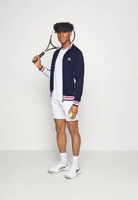 Marineblå lynlåsjakke med røde og hvide accenter, hvid T-shirt, hvide shorts og hvide sko, der holder en tennisketcher og en bold.