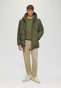 Grüne Pufferjacke mit Reißverschlusstaschen, kombiniert mit einem grünen Pullover und beigen Hosen. Weiße und beige Sneaker vervollständigen das Outfit.