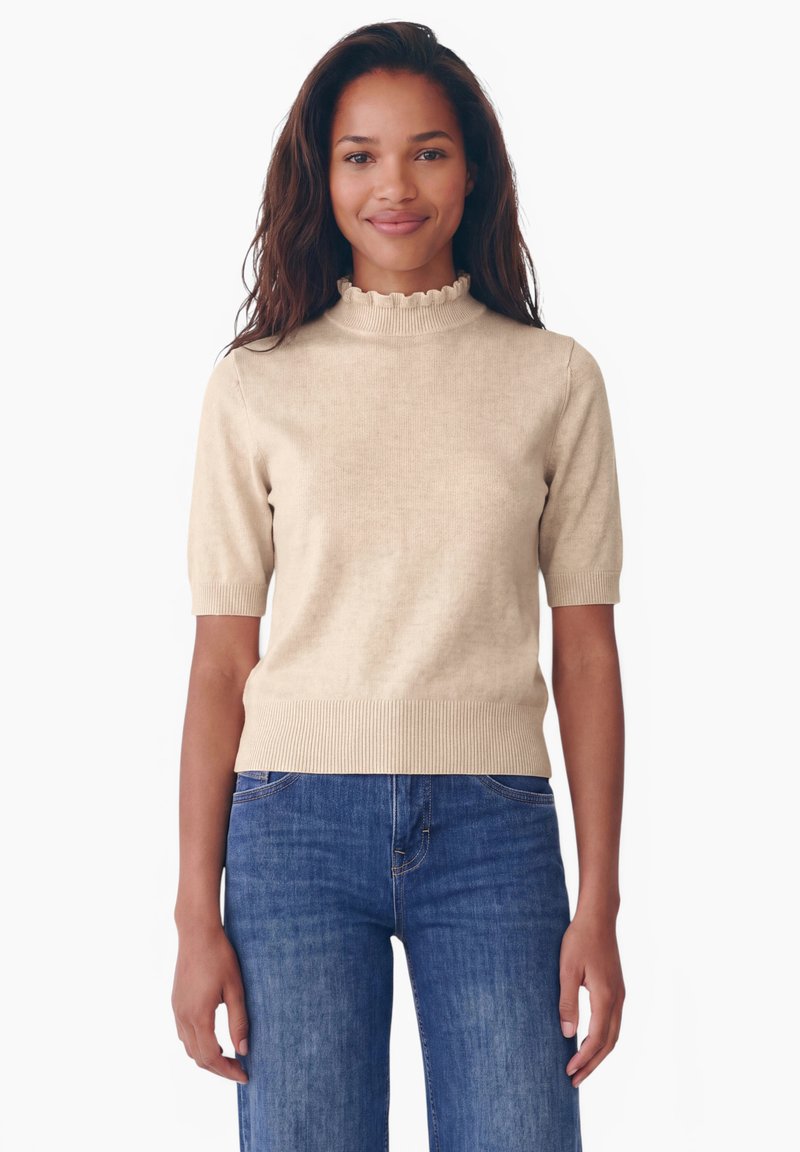 Femme aux longs cheveux foncés portant un pull beige à manches courtes avec un col à volants et un jean bleu, debout et souriante sur un fond blanc.