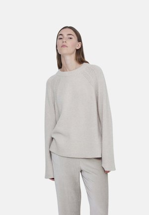 Joven mujer con suéter de punto beige claro y pantalones acanalados a juego, de pie frente a un fondo liso.