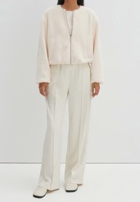 Chaqueta difusa y recortada en rosa suave, con cremallera delantera y dos bolsillos, combinada con pantalones anchos blancos y zapatos color crema.