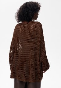 Brązowy dzianinowy poncho o otwartym splotie, z długimi rękawami i luźnym krojem. Posiada okrągły dekolt oraz szczegóły wykończenia w żebrowanym stylu.