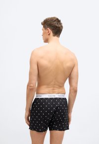 Νεαρός άνδρας, φαινομενικά από πίσω, φοράει μαύρα μποξεράκια Calvin Klein με άσπρη ζώνη μέσης με λογότυπο, σε απλό φωτεινό φόντο.