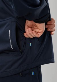 Personne ajustant le poignet d'une veste bleu marine avec une poche zippée et une petite étiquette avec le drapeau suédois près de l'ourlet.